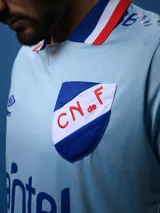 Camiseta de fútbol celeste con cuello y mangas con detalles en azul y rojo. Incluye logo de Umbro, escudo del Club Nacional de Football y sponsor Antel.