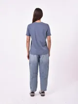 Remera blanca de algodón con cuello redondo y manga corta. Presenta un pequeño bordado en el pecho izquierdo con una figura que parece ser una persona acostada en una tabla de surf o flotador.