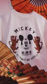 Remera blanca de algodón con cuello redondo y mangas cortas. Estampado frontal con diseño de Mickey Mouse, tigres y texto en inglés y chino.