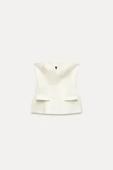 Top blanco de escote recto con detalle de pieza metálica en forma de V forrada y hombros descubiertos. Tiene falsos bolsillos con solapa en el delantero, forro interior combinado y cierre lateral con cremallera oculta en la costura.