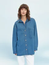 Camisa de jean celeste, de corte amplio, con cuello clásico, cierre frontal con botones y bolsillo de parche en el pecho.