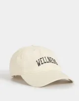 Gorra deportiva color crudo, de tela canvas con bordado frontal de la palabra "Wellness" en letras negras estilo universitario. Tiene visera curva y tira ajustable trasera con hebilla metálica.