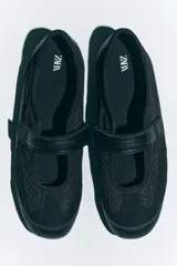 Zapato tipo bailarina color negro, con capellada de cuero sintético y rejilla. Tiene tira con velcro en el empeine y suela plana.
