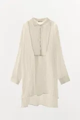 Camisa color crudo de corte holgado, confeccionada en hilatura con lino y tejido con efecto arrugado. Tiene cuello solapa, mangas largas con puños, bajo asimétrico con aberturas laterales y cierre frontal con botones.