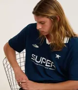 Remera de manga corta azul marino con cuello polo blanco. Presenta un estampado frontal en blanco con la palabra "SUPER" y un texto más pequeño debajo, junto con una estrella y un logo de platillo volante.