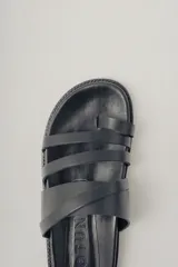 Sandalia plana negra de cuero con diseño de tiras anchas entrecruzadas sobre el empeine y una tira que separa el dedo gordo. Suela plana y gruesa.