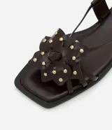 Sandalia plana color marrón con punta cuadrada, tiras finas con flor central y strass, y cierre de tira fina con hebilla cuadrada.