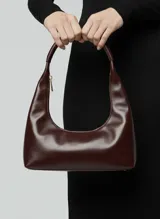 Bolso de hombro de efecto cuero color marrón oscuro (Port Royale), con forma de media luna y asa única. Presenta un acabado brillante y un tirador de cremallera metálico dorado.