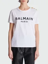 Remera blanca de algodón con cuello redondo y mangas cortas. Estampado frontal con el logo 'BALMAIN PARIS' en color negro. Detalle de botones dorados en el hombro izquierdo.