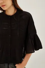 Camisa blanca de algodón con cuello alto y volados, mangas largas abullonadas con puños elásticos. Presenta detalles de encaje y frunces en el frente y la espalda.