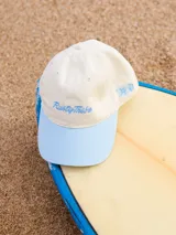 Gorra de lona color crema con visera celeste y bordado frontal con el logo de la marca Rusty en celeste.