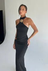 Vestido largo negro ajustado al cuerpo, con escote halter que se une a un detalle metálico dorado en el centro del pecho. Presenta frunces drapeados en la zona de la cadera y un corte asimétrico en el lateral.
