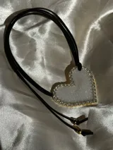 Collar con dije de corazón blanco brillante con borde dorado y strass, colgando de cordón negro ajustable.