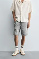Bermuda de denim de algodón, color gris lavado, con corte relaxed fit y roturas visibles en la parte delantera de ambas piernas. Presenta cinco bolsillos y cierre frontal con cremallera y botón.
