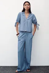 Camisa celeste de denim con cuello solapa, escote en pico, mangas abullonadas con lazos y cierre frontal con botones.