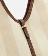 Bolso tipo tote de lona con rayas verticales en tonos beige y marrón claro. Presenta ribetes y asas de cuero sintético marrón oscuro con costuras contrastantes. Tiene un cierre central con tira de ajuste y hebilla metálica dorada.