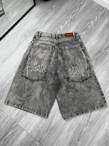 Bermuda de jean gris oscuro con efecto lavado, corte ancho y tiro alto. Presenta bordados blancos en estilo gótico o tribal en los bolsillos traseros. Tiene un parche de cuero en la cintura con la inscripción "JAQUEMATE".