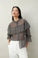 Camisa negra semitransparente de encaje bordado con diseño geométrico. Tiene silueta fluida, mangas largas y volados en el frente y los hombros.