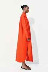 Vestido camisero largo color naranja, de corte amplio, con cuello clásico, cierre frontal con botones, mangas largas con puños abotonados, bolsillos laterales y tajos laterales pronunciados.