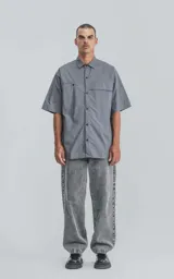 Camisa gris de manga corta confeccionada en poplin 100% algodón, con silueta oversize y dos bolsillos delanteros, uno con solapa y otro con ojal y botón.