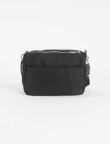 Bandolera negra de tela con múltiples cierres horizontales plateados. Incluye una correa ajustable y detalles colgantes en los tiradores de los cierres, uno de ellos es una borla trenzada blanca y negra y otro es un tirador pequeño de color amarillo.