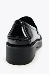 Zapato tipo mocasín de charol negro, con detalle de antifaz en el empeine y punta redonda.