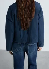Chaqueta azul de estilo tejano vaquero con diseño acolchado y relleno. Tiene cuello redondo, cierre de cremallera frontal, dos bolsillos de ribete delanteros y mangas largas.