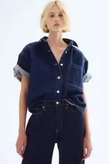 Camisa azul de denim, corte amplio, con cuello clásico, abotonadura frontal, manga corta, bolsillo de parche en el pecho y canesú en la espalda.