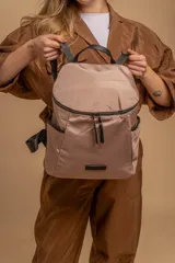 Mochila urbana de diseño compacto, color beige claro con acabado satinado. Presenta un bolsillo frontal con cierre doble y bolsillos laterales. Las correas y detalles son de color negro.