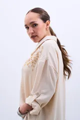 Camisa color beige con bordado de lentejuelas doradas en los hombros, corte clásico, cuello camisero y mangas largas con puños abotonados.