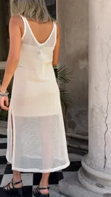 Vestido largo de tejido de red blanco, con apliques de flores negras en relieve. Lleva tirantes finos y tiene un corte recto con abertura lateral. Se usa sobre una prenda interior blanca.