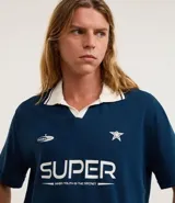 Remera de manga corta azul marino con cuello polo blanco. Presenta un estampado frontal en blanco con la palabra "SUPER" y un texto más pequeño debajo, junto con una estrella y un logo de platillo volante.