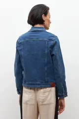 Chaqueta de jean azul medio, de corte clásico, con cuello camisero, bolsillos con solapa en el pecho y cierre frontal con botones metálicos color cobre.