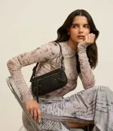 Cartera tipo baguette de PU negro, con acabado liso y detalles de herrajes metálicos en tono gunmetal, incluyendo remaches y un anillo decorativo en el frente. Posee un bolsillo frontal con cierre y un asa corta ajustable.