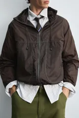 Cazadora de tejido técnico ripstop color marrón oscuro, con cuello alto y capucha ajustable, mangas largas con puños elásticos, bolsillos frontales con cremallera oculta, bajo ajustable con elástico lateral y cierre frontal con cremallera.