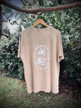 Remera oversize de manga corta, color beige lavado, con estampado circular en la espalda que incluye una palmera y texto que dice "SUNSET", "PALM", "TROPICAL" y "BEACH TRIP".