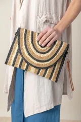 Sobre de mano de rafia con diseño de rayas horizontales en tonos beige, dorado y negro. Tiene cierre superior y correa de cuero sintético marrón.