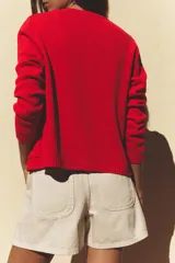 Chaqueta roja de punto con cuello redondo y manga larga. Tiene bolsillos delanteros de plastrón, cierre frontal con botones dorados con relieve y acabados en rib.