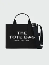 Bolso tote negro de lona con doble asa de mano y correa bandolera ajustable y extraíble. Estampado frontal con el texto "THE TOTE BAG MARC JACOBS" en blanco.