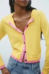 Chaqueta de punto amarilla con cuello redondo, manga larga y cierre frontal con botones negros. Presenta ribetes rosas en cuello, puños y bajo.