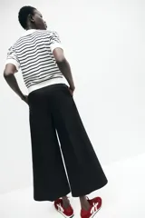 Pantalón palazzo negro de tiro alto con elástico oculto en la cintura, cierre lateral oculto y pinzas que realzan la figura. Modelo de corte holgado con bolsillos al bies, bolsillos decorativos atrás y piernas tres cuartos con raya adelante y atrás.