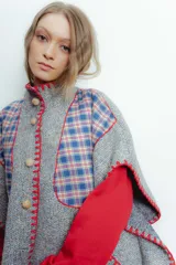 Poncho de paño gris con apliques de tela escocesa y bordado artesanal en color rojo. Tiene cuello alto, cierre con botones de madera y mangas 3/4.