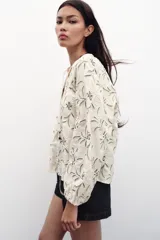 Blusa color crema con bordado floral en azul, de cuello redondo con cordón ajustable y mangas largas abullonadas con puños elásticos.