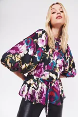 Camisa estampada con flores en tonos morados, azules, amarillos y verdes sobre fondo negro. Tiene cuello redondo, mangas abullonadas 3/4 y lazo en la cintura.