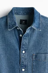 Camisa de denim azul de algodón rígido, corte holgado, cuello inglés, tapeta clásica con botones blancos, bolsillo de parche en el pecho, canesú en la espalda, mangas largas con puños abotonados y bajo levemente redondeado.