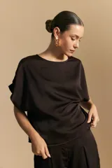 Top negro de manga corta con silueta amplia y caída fluida, confeccionado en un tejido liviano con leve brillo. Presenta un botón de nácar en la espalda con una pequeña abertura y mangas amplias tipo kimono.