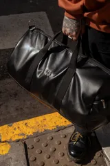 Bolso duffel negro de cuero sintético con logo de ancla bordado en rosa. Tiene doble asa de mano reforzada, correa de hombro regulable y desmontable, cierre superior con doble cremallera y tres bolsillos (dos exteriores y uno interior).