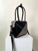 Cartera rígida negra de cuero con tachas plateadas y tres combinaciones de cuero diferentes que aportan textura. Tiene herrajes y cierre metálico plateado marca YKK. Cuenta con un asa fina para llevar al hombro o en la mano y una correa larga extra regulable.