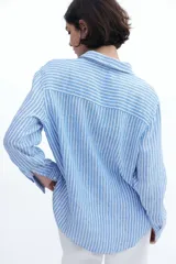 Camisa celeste y blanca a rayas verticales, de lino liviano, con cuello clásico, botones frontales, canesú de doble capa en la espalda, hombros caídos, mangas largas con puños abotonados, bolsillo de parche en el pecho y bajo redondeado.