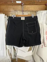 Short de baño negro de largo medio (18 pulgadas) con costuras reforzadas en hilo blanco contrastante. Presenta cordón de ajuste en la cintura y un pequeño logo estampado en la pierna derecha.